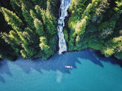 Aerial view of a serene waterfall cascading into a turquoise lake, surrounded by lush green trees and a small boat.