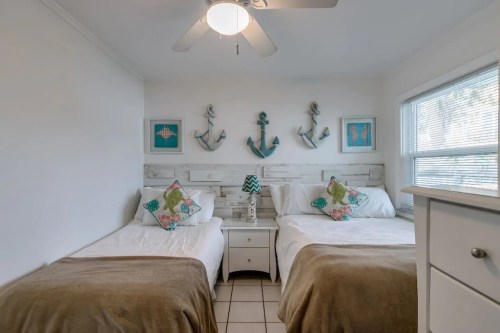 Cozy bedroom with two twin beds, nautical decor, and a ceiling fan, featuring light colors and decorative pillows.