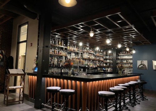 A stylish bar with a wooden counter, high stools, and shelves filled with various bottles, illuminated by warm lights.