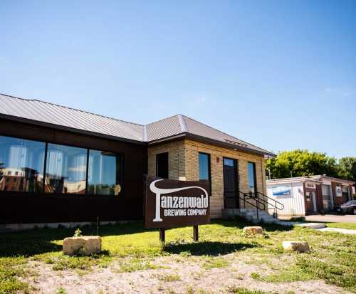 Exterior of Tanzanwald Brewing Company, featuring a brick building and a sign in a grassy area under a clear blue sky.