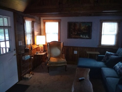 Cozy cabin interior with a yellow armchair, blue sofa, lamp, and wooden decor, featuring windows and a door.