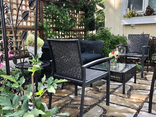 A cozy outdoor seating area with black wicker furniture, a glass table, and lush greenery in a garden setting.