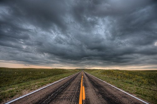 A long, empty road stretches into the distance under a dramatic, cloudy sky. Lush green fields line the sides.