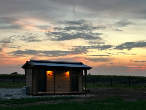 A small wooden cabin illuminated at dusk, with a colorful sky and fields in the background.