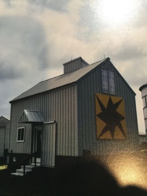 A gray barn with a metal roof, featuring a large yellow star on the side and a small porch at the entrance.