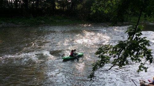 A person in a green kayak paddles through a river with gentle waves, surrounded by trees and sunlight.