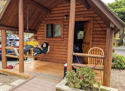 A person relaxes on a swing outside a wooden cabin, surrounded by greenery and outdoor furniture.