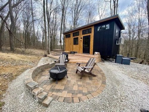 A cozy tiny house with a wooden exterior, surrounded by trees and a stone patio with chairs and a fire pit.