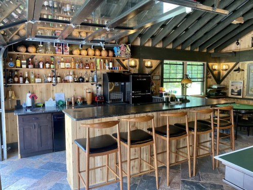 A stylish bar area with wooden stools, shelves of liquor, and a coffee machine in a rustic setting.