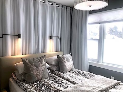 Cozy bedroom with a double bed, decorative pillows, soft lighting, and sheer curtains by a window.
