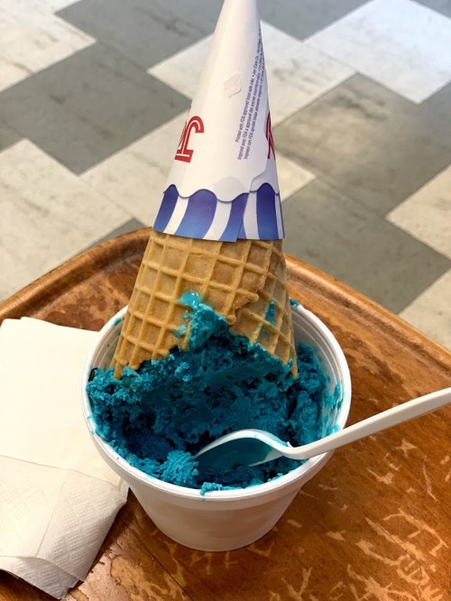 A cup of bright blue ice cream with a waffle cone on top, resting on a wooden tray with a napkin and spoon.