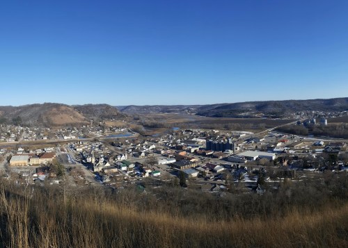 A panoramic view of a small town nestled in a valley, surrounded by hills and clear blue skies.