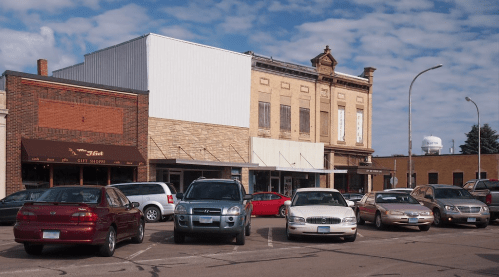 A small town street scene featuring a mix of parked cars and historic buildings under a partly cloudy sky.