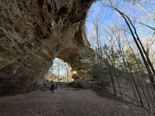 A large rock arch in a forested area, with two people walking on sandy ground beneath it.