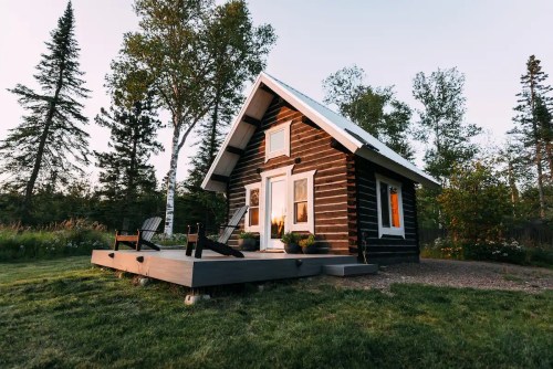 A cozy log cabin with a deck, surrounded by trees and greenery, during a serene sunset.