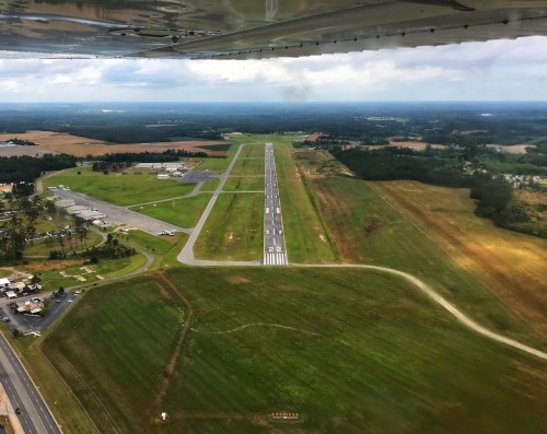 Aerial view of an airport runway surrounded by green fields and scattered buildings under a cloudy sky.