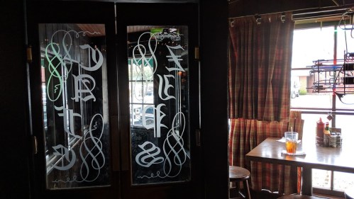 A decorative glass door with intricate white lettering, leading to a cozy dining area with a table and drink.