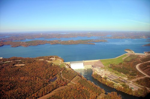 Aerial view of a dam surrounded by lush greenery and a large lake, with hills and islands in the background.