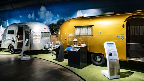 Two vintage trailers, one silver and one yellow, displayed in a museum setting with informational exhibits nearby.
