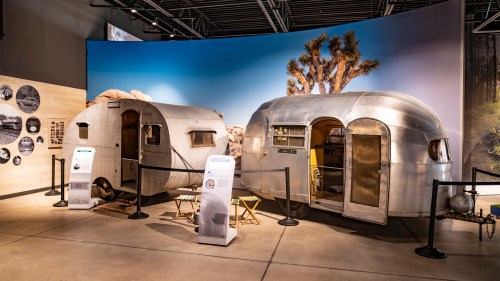 Two vintage trailers on display in a museum, with informational panels and a backdrop of a desert landscape.