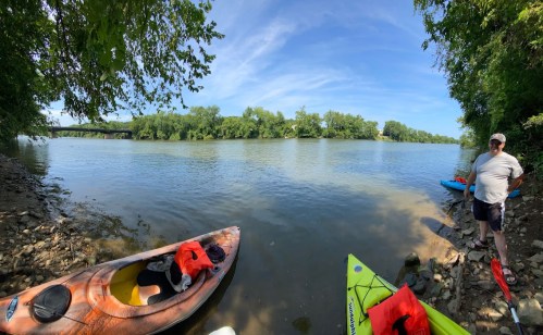 A serene river scene with kayaks on the shore, surrounded by lush greenery and a clear blue sky.