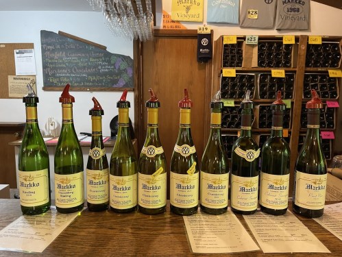 A row of wine bottles with yellow labels on a wooden counter, alongside menus and a chalkboard in the background.
