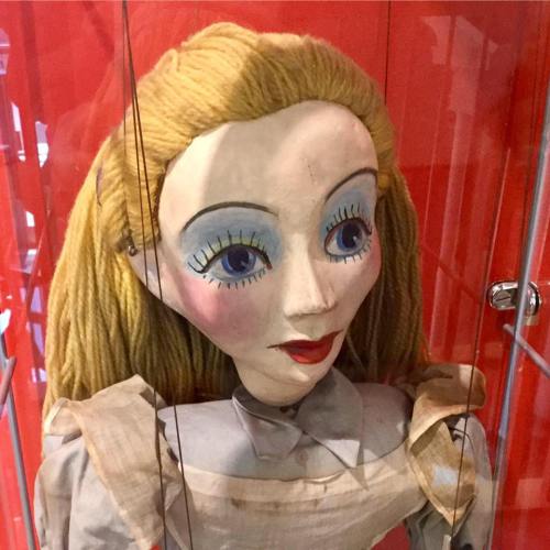 A vintage puppet with blonde hair and blue eyes, displayed in a glass case against a red background.