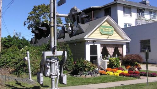 A quaint yellow building with a sign reading "Millenhurst Station" next to a railroad crossing and colorful flower beds.
