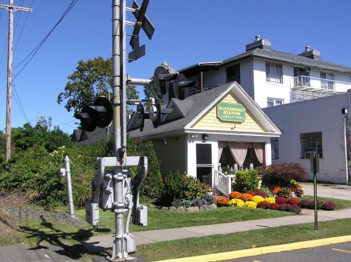 A quaint building with a sign reading "Allenhurst Station" near a railroad crossing, surrounded by colorful flowers.