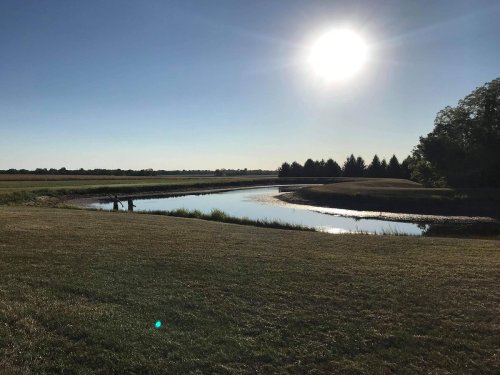 A serene landscape featuring a calm river, green fields, and a bright sun in a clear blue sky.