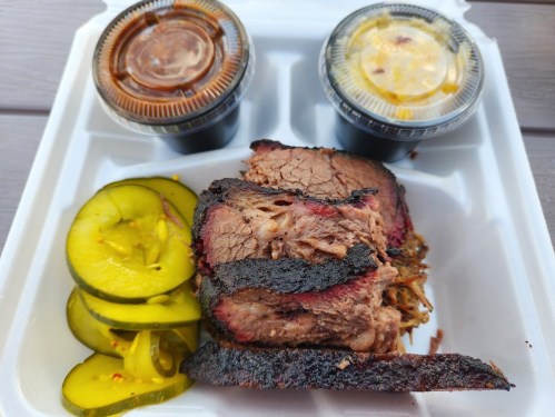 A serving of sliced brisket with pickles and two cups of barbecue sauce on a takeout container.