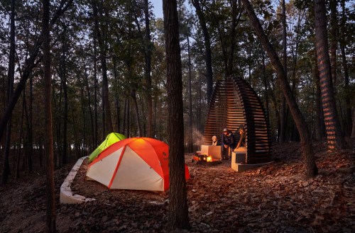 A campsite in the woods at dusk, featuring colorful tents and a cozy fire pit surrounded by trees.