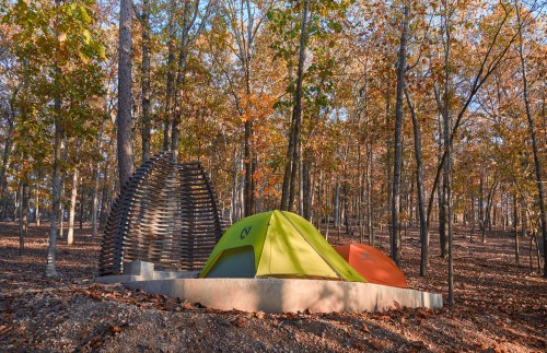 A green and orange tent set in a wooded area with autumn leaves and a unique wooden structure nearby.