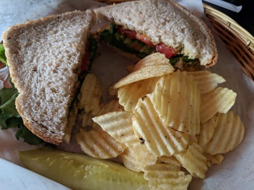 A sandwich with lettuce and tomato, served with ridged potato chips and a pickle on the side, in a basket.