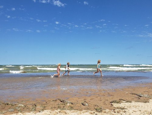 Three children play and run along the shoreline, with waves gently crashing in the background under a clear blue sky.