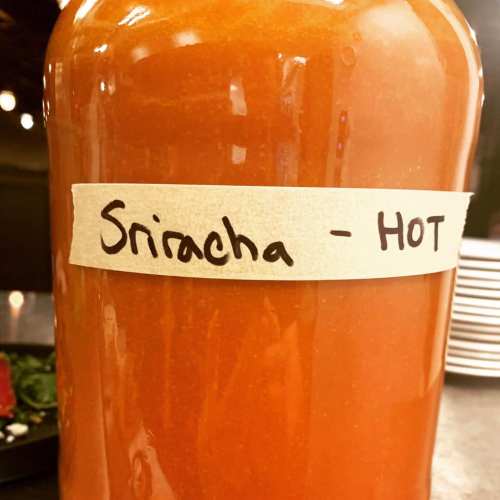 A glass jar filled with red sriracha sauce, labeled "Sriracha - HOT" on a paper tag.