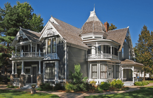 A large, ornate Victorian-style house with a turret, intricate woodwork, and a lush garden under a clear blue sky.