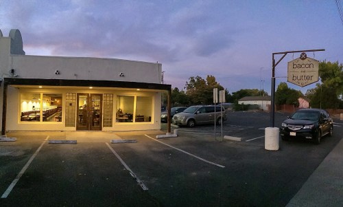 Exterior of a restaurant named "Bacon Butter" at dusk, with parked cars and a sign visible.
