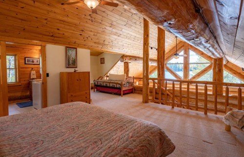 Cozy log cabin interior featuring a bedroom with a quilt, wooden beams, and large windows overlooking nature.
