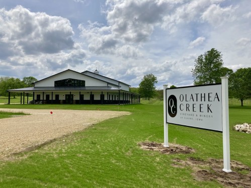 A modern winery building with a sign for Olathea Creek Vineyard & Winery, surrounded by green grass and trees.