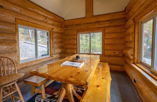 Cozy log cabin interior featuring a wooden dining table, chairs, and large windows with a view of nature.