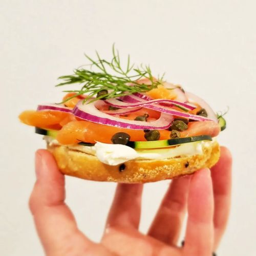 A hand holding a bagel topped with cream cheese, smoked salmon, cucumber, red onion, capers, and fresh dill.