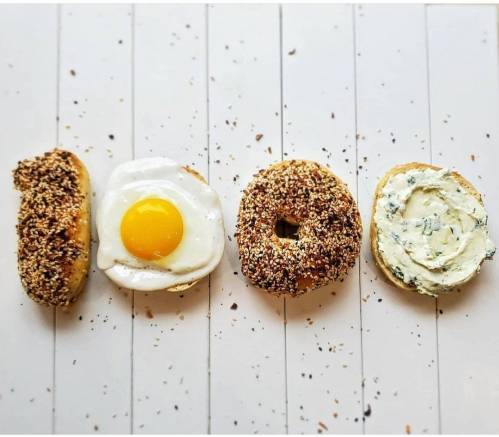 Four bagels arranged to spell "100," topped with seeds, a fried egg, and cream cheese.