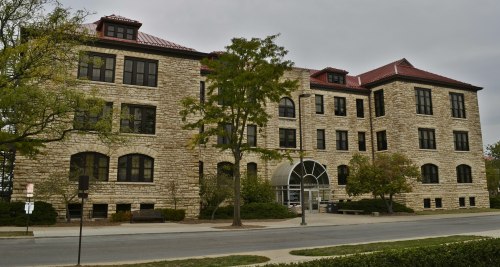 Historic stone building with large windows and a red roof, surrounded by trees and a sidewalk. Overcast sky.