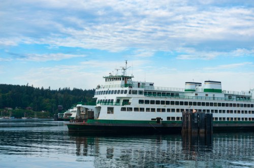 A green and white ferry docked at a pier, reflecting in calm water under a cloudy sky.