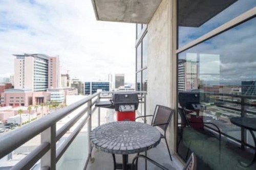A balcony with a round table and chairs, featuring a grill, overlooking a city skyline with buildings and clouds.