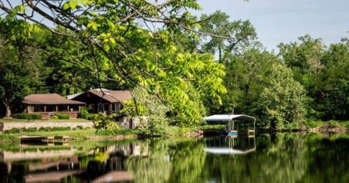 A serene lakeside view featuring a house, dock, and lush green trees reflecting in the calm water.