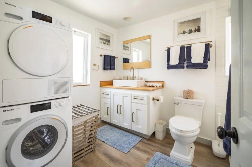 A bright, modern bathroom featuring a stacked washer and dryer, a sink with a mirror, and a toilet, all in a clean design.