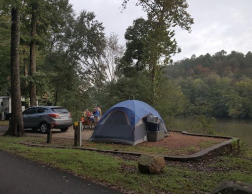 A blue tent by a river, with a car parked nearby and people preparing food at a picnic table under trees.