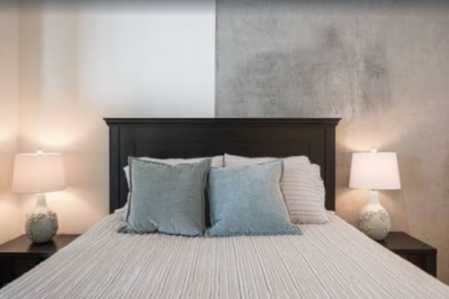 A modern bedroom featuring a bed with light blue pillows, two bedside lamps, and a concrete wall backdrop.